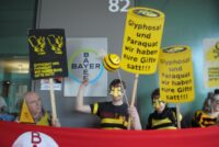 Protestierende stehen mit Parolenschilder vor dem Hauptsitz von Bayer in Basel. Das Logo von Bayer ist im Hintergrund zu erkennen. Eine Person zeigt den "Vogel". Im Vordergrund ist das rote Banner 