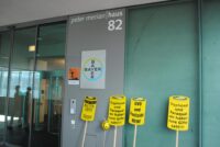 Der Eingang von Bayer mit Logo und Warnhinweis "Umweltgefährlich" neben dem Eingang. An die Wand lehnen Protestschilder und eine Biene aus Papier-mâché
